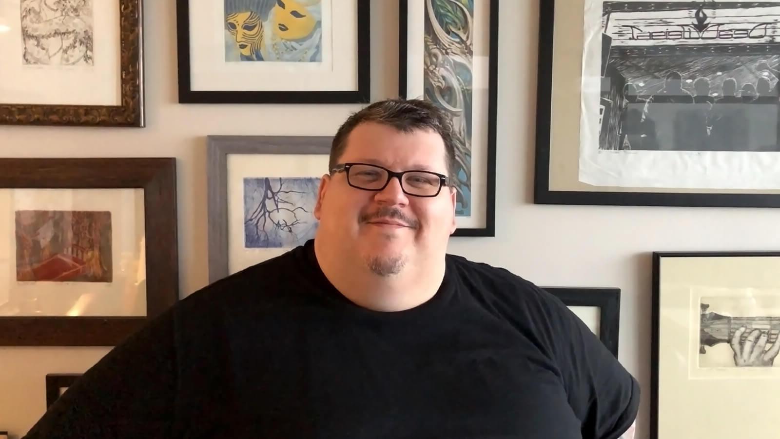 一个面带微笑的男人戴着眼镜，穿着黑色t恤，坐在墙上挂满了镶框的画作.