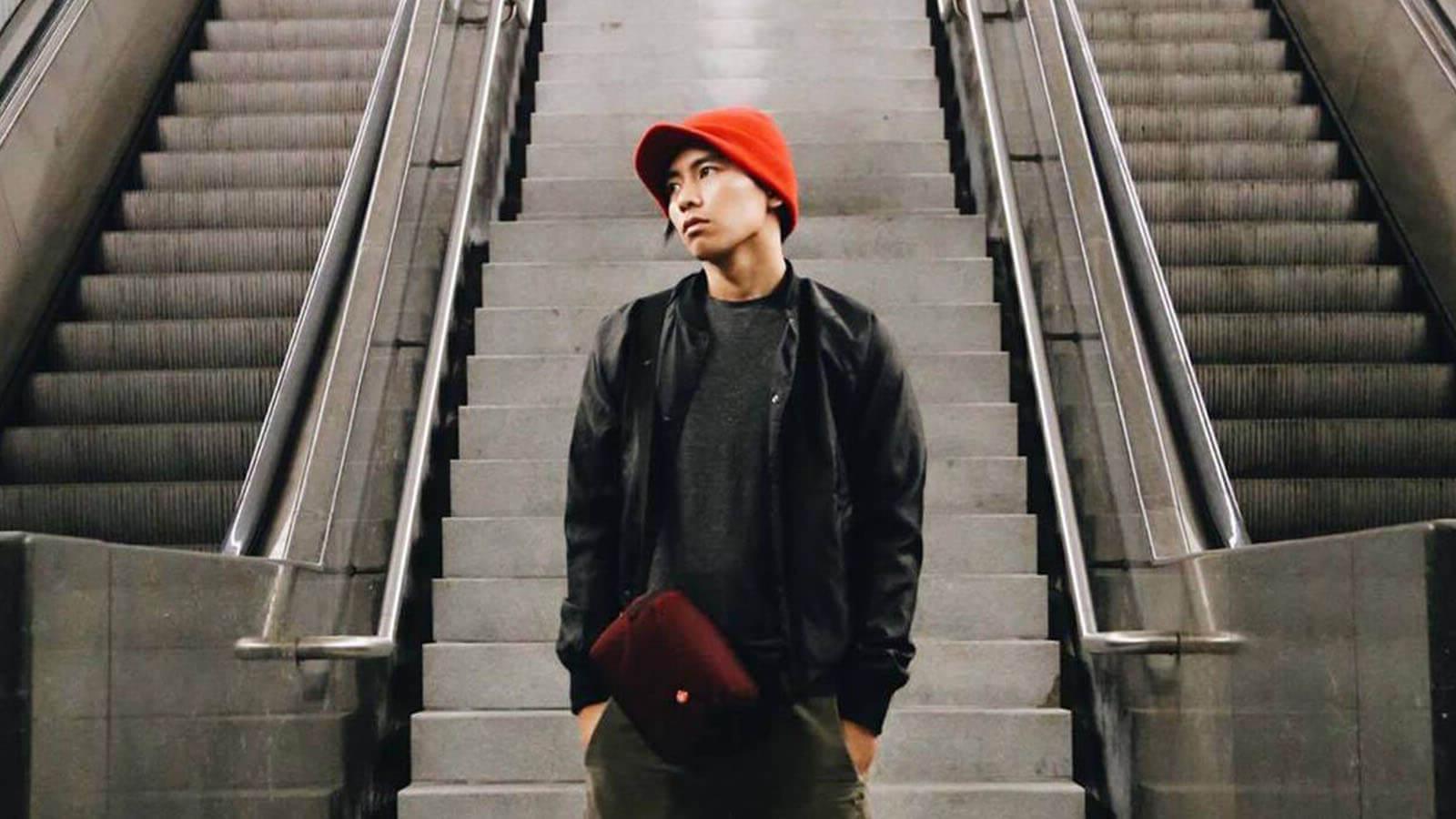 A young Asian man wearing a red knit cap, 黑色夹克和衬衫, 绿色的裤子, and a burgundy bag stands between two escalators. His hands are in his pockets and he is looking off to the left.
