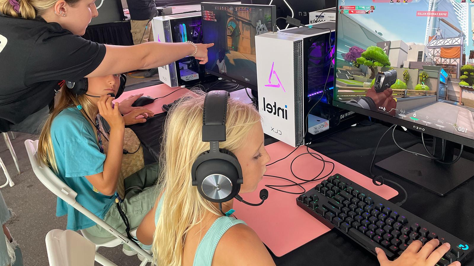 两个小女孩坐在台式电脑前玩《我的世界》(Minecraft)，一个穿着超级女声玩家专业版衬衫的女人在旁边帮助她们.