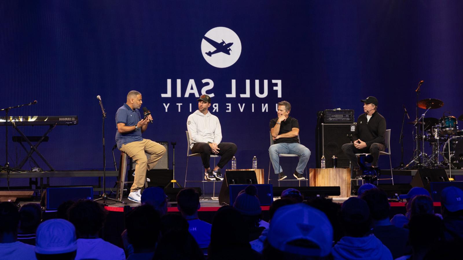 MLB Pitcher Adam Wainwright and three other people are seated on a stage during a panel, the large-scale LED screen behind them reads “满帆大学” against a blue backdrop.