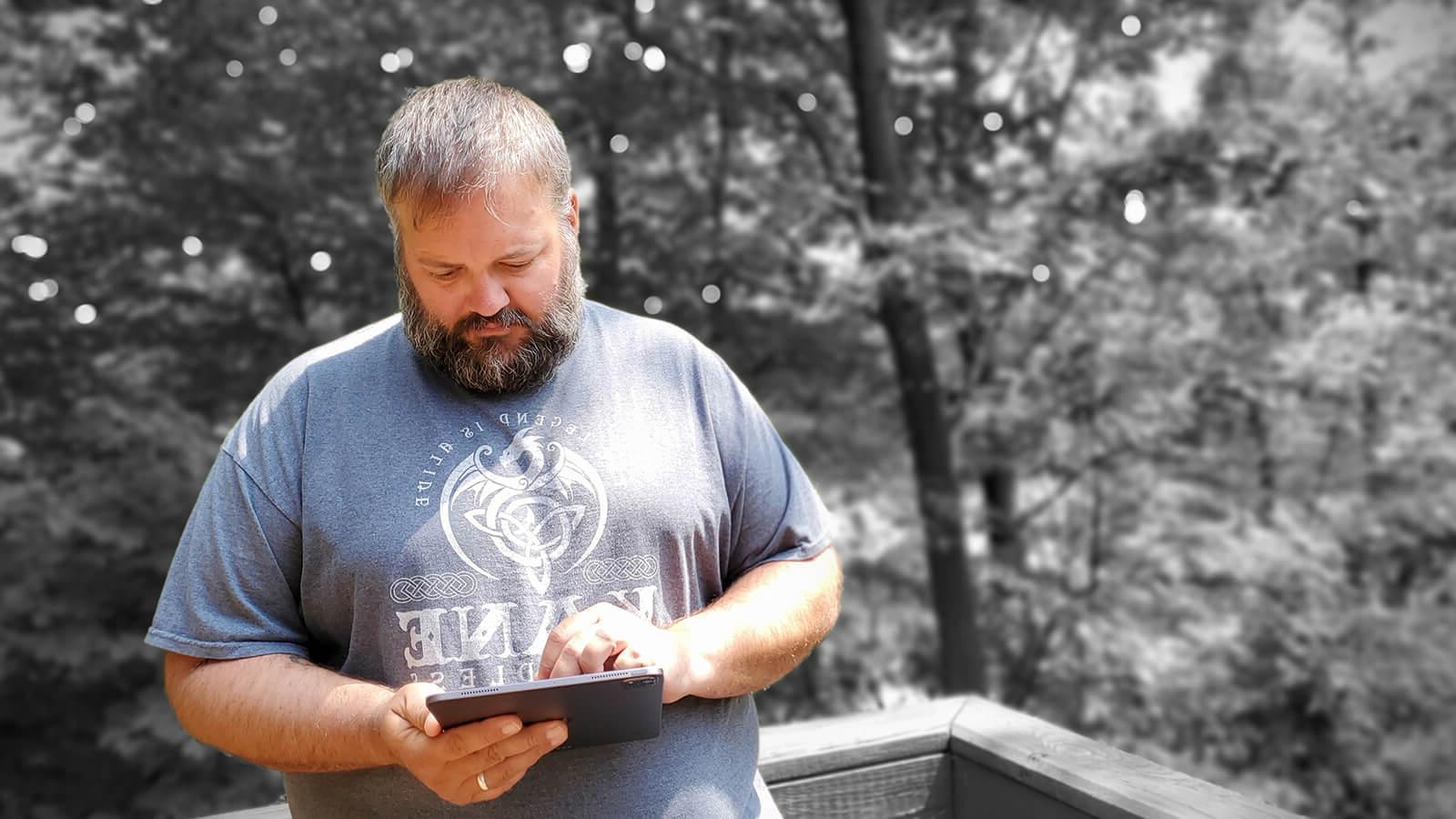 Grad Ryan Backa in a blue shirt with a white dragon design, holding an electronic tablet in front of a wooded area in greyscale.