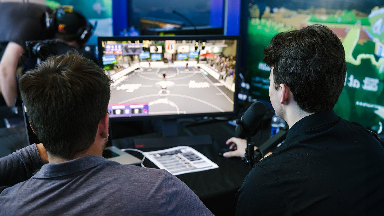 两个人背对着镜头，在他们面前桌子上的一个小显示器上观看“NBA 2K”游戏.