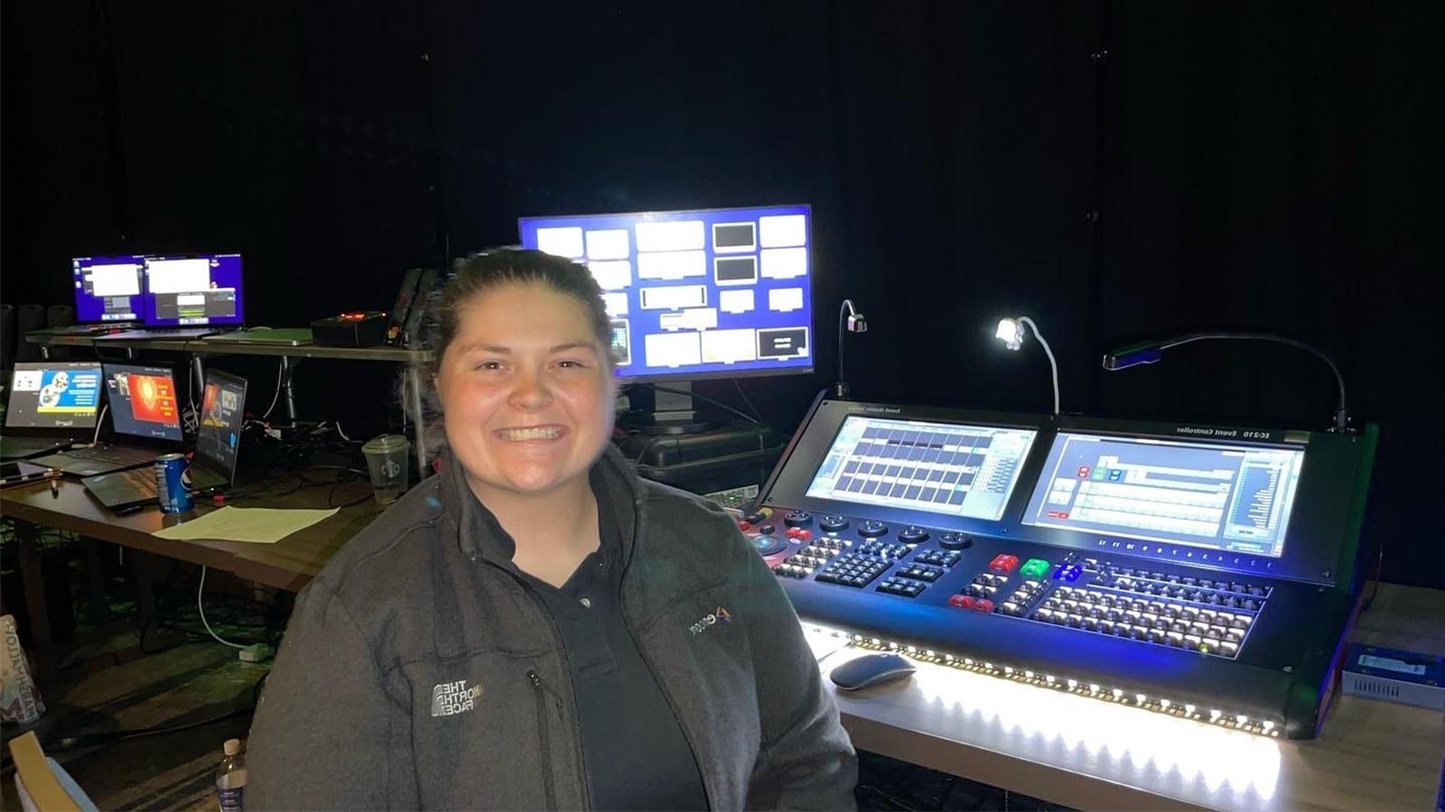 Jean Fuemmeler sits in front of live event video production controllers. She is wearing a gray jacket and smiling.