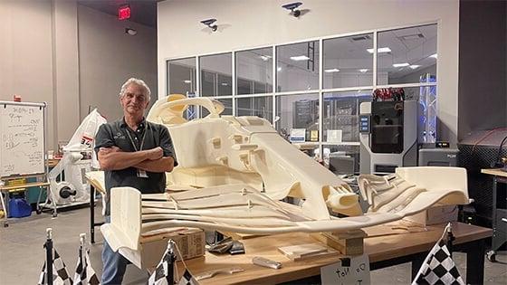 课程主任帕特·斯塔雷斯, 一个白发男子，身穿灰色polo衫, standing next to a large workbench with a 3D-printed replica of a Formula 1 race car in 满帆’s SimLab.