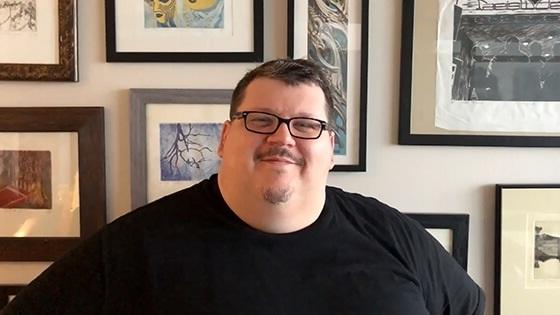 一个面带微笑的男人戴着眼镜，穿着黑色t恤，坐在墙上挂满了镶框的画作.