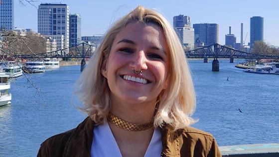 毕业生Angela Decastro, a woman with shoulder-length blonde hair wearing a brown jacket and white top, 站在俯瞰河流的桥上微笑, 在她身后的远处是一座桥和城市景观.