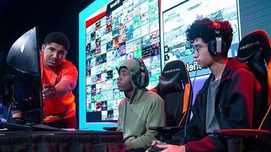 Two Florida high school students seated on stage in front of a gaming monitor with a Full Sail Armada athlete helping them to their right.
