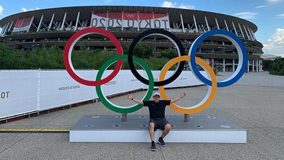 Grad Andrew Molina sits outdoors with his arms spread in front of a set of Olympic rings. There is a stadium with a banner for the 2020 Tokyo Olympics behind him.