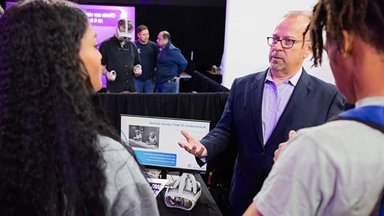 Man wearing glasses and a blue suit speaks to guests at Career Fair while man in back tries on Virtual Reality goggles.