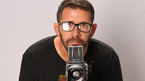 塞巴斯蒂安·克里斯蹲在摄像机后面. 他戴着一副黑框眼镜，穿着一件印有商标的黑色t恤.