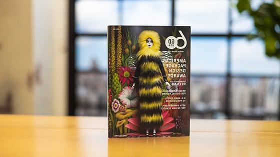 Graphic 设计 USA’s April 2023 issue sits upright on a wooden table. The cover features a female doll wearing a long furry black-and-yellow dress with a hood. 她站在一个迷幻的花朵背景前.