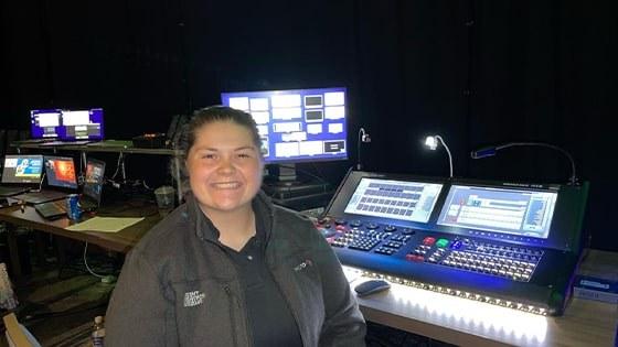 Jean Fuemmeler sits in front of live event video production controllers. 她穿着一件灰色的夹克，面带微笑.