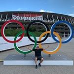 毕业生安德鲁·莫利纳坐在户外，双臂摊开，面前是一组奥运五环. 他身后是一个体育场，上面挂着2020年东京奥运会的横幅.