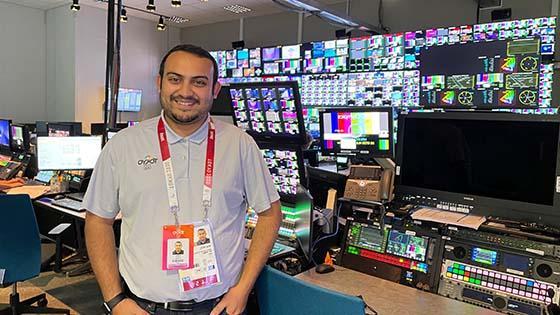 Niraj Patel站在一个满是电视屏幕的广播室里. 他穿着一件灰色的纽扣衬衫，胸前有2020年东京奥运会的标志.