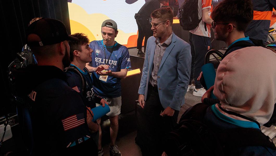 A man in a light blue sport coat stands near a stage while talking to a group wearing Butler Esports jerseys.